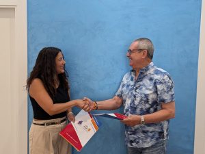 Una mujer con un top negro y pantalón beige sonríe mientras estrecha la mano de un hombre con camisa azul estampada y gafas. Ambos sostienen las carpetas rojas y blancas del convenio con Novaluz Energia, la ACET y el CCA Torremolinos Abierto.
